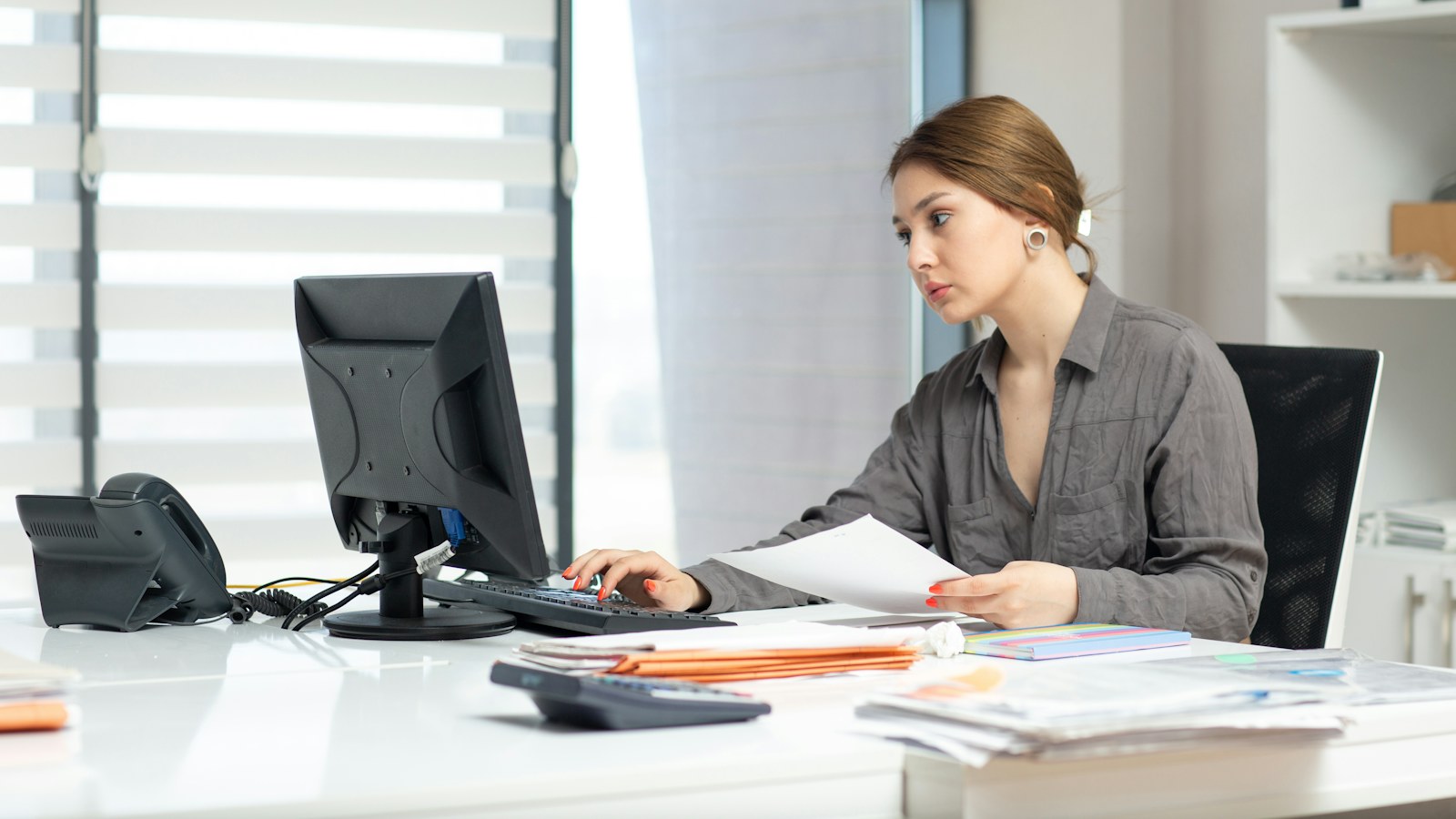 An accounts receivable clerk typing a collection email at an office keyboard while holding a printed invoice, with a desk phone and stacks of paperwork beside her
