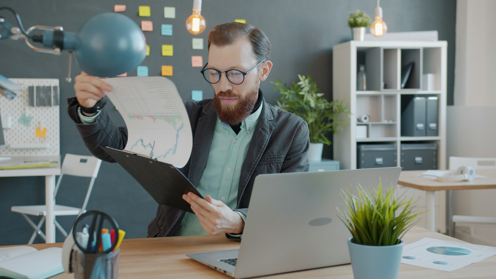 A bearded credit manager in glasses and a dark blazer lifting a printed aging report off a clipboard beside his laptop, checking how long an unpaid commercial invoice has been outstanding